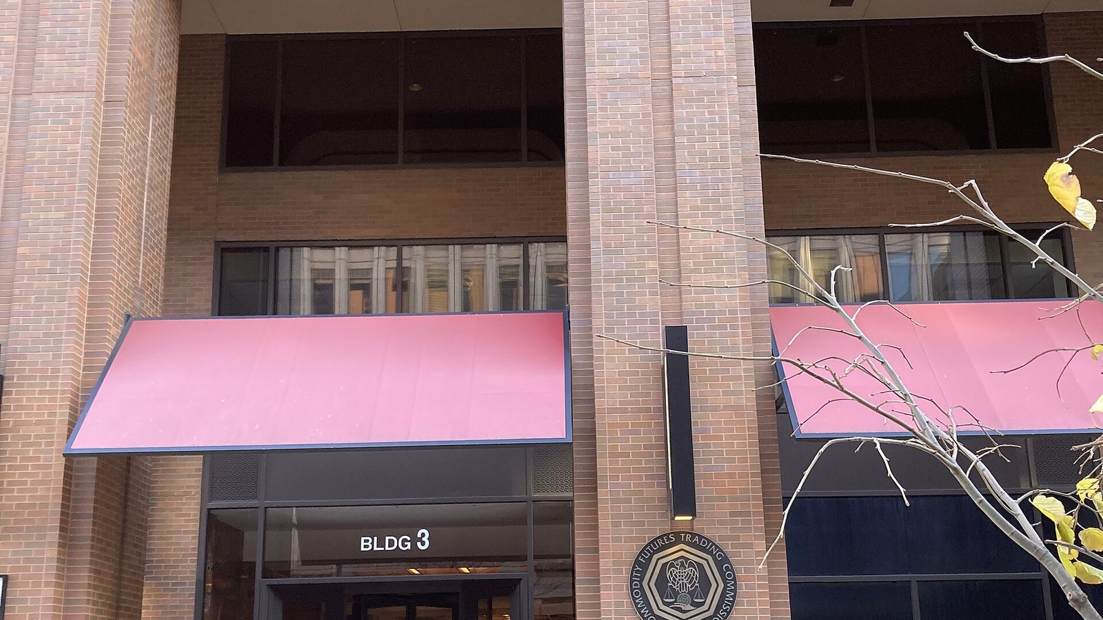 The entrance to the Commodity Futures Trading Commission headquarters at Lafayette Centre in Washington: red brick piers, two pink awnings over glazed doors marked BLDG 3, and the Commission seal mounted on the brickwork beside them, with bare winter branches at the right of the frame.
