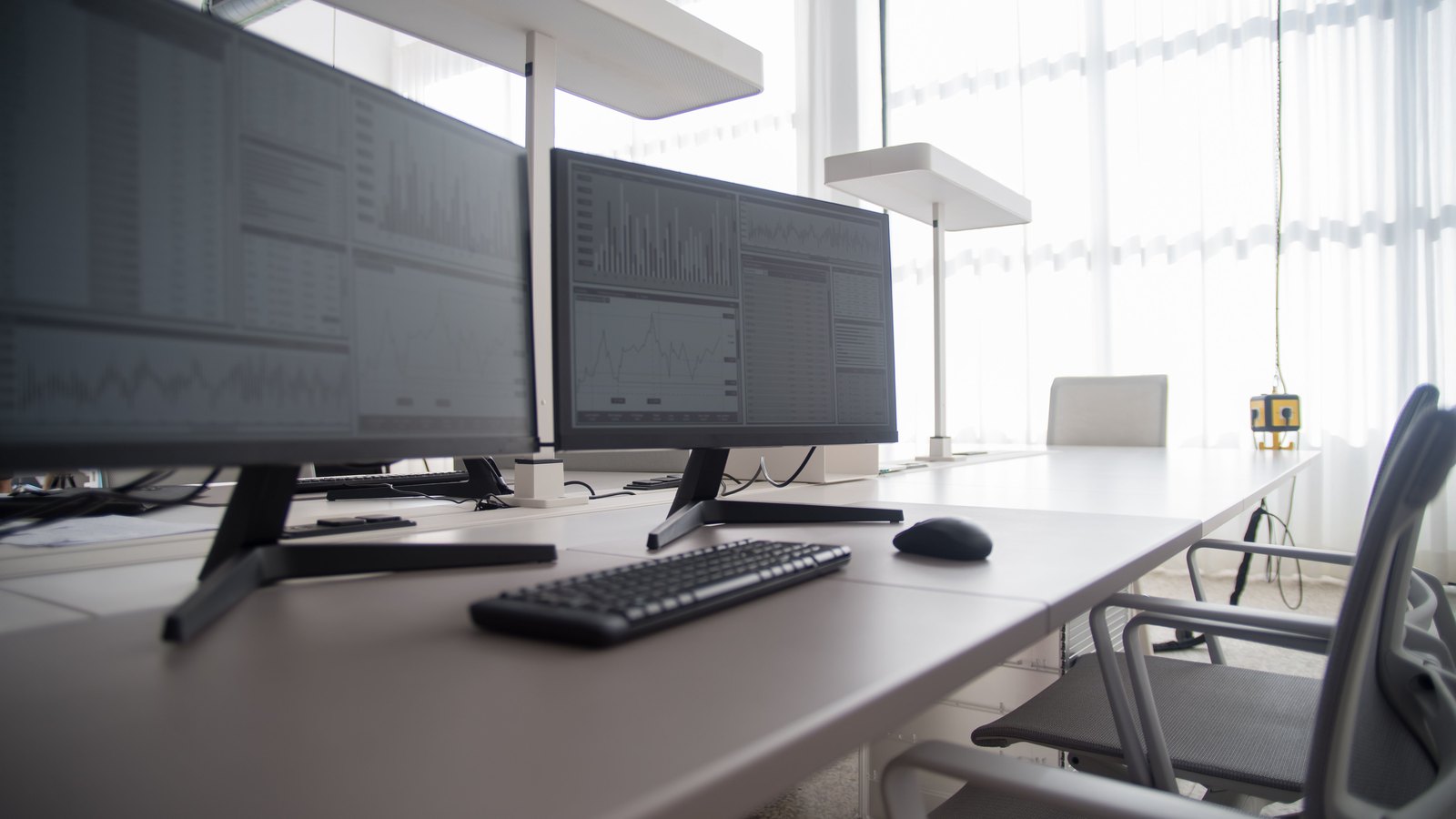 An empty white office desk carrying two large monitors, each showing grey charts and dense tables of figures too small to read. A keyboard, a mouse and an empty chair sit in front of them, with a bright curtained window behind.