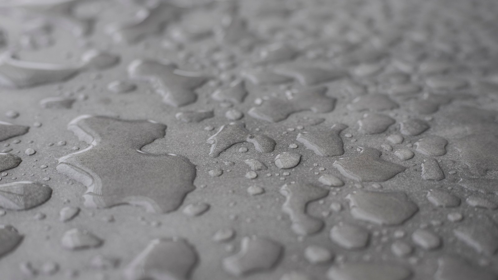 A close photograph of scattered water droplets beaded on a flat matte grey surface. The larger drops have flattened into irregular pools with bright rims, the smaller ones still hold round, and the surface falls gently out of focus toward the top of the frame. No text, markings, brand marks, people or machinery are in view. A generic material close-up, not a photograph of any ChargePoint site, vehicle or charging equipment.