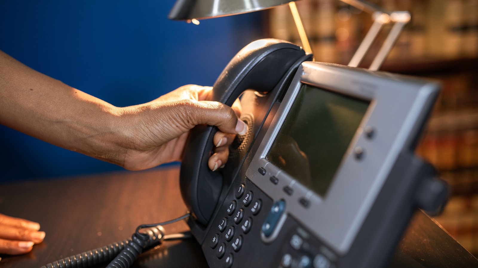 A close photograph of a hand lifting the black corded handset from a grey desk telephone standing on a polished wooden counter. The phone has a small blank screen, a row of soft keys and a numeric keypad, and its coiled cord runs off to the left. A deep blue wall and a lit desk lamp are behind it, with shelves of books out of focus on the right. No text, signage or identifying detail is legible. A generic illustration, not a photograph of any Charlotte County facility.