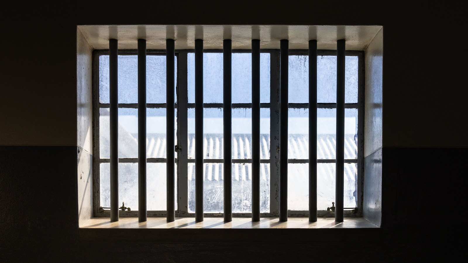 A barred window set in a dark concrete wall, daylight coming through the gaps and falling across the sill. Stock photo, not a photograph of this event.