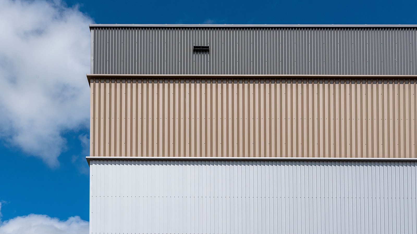 The upper corner of a large industrial building clad in vertical corrugated metal, in three horizontal bands of grey, tan and white, photographed straight on against a deep blue sky with a white cloud at the left edge.