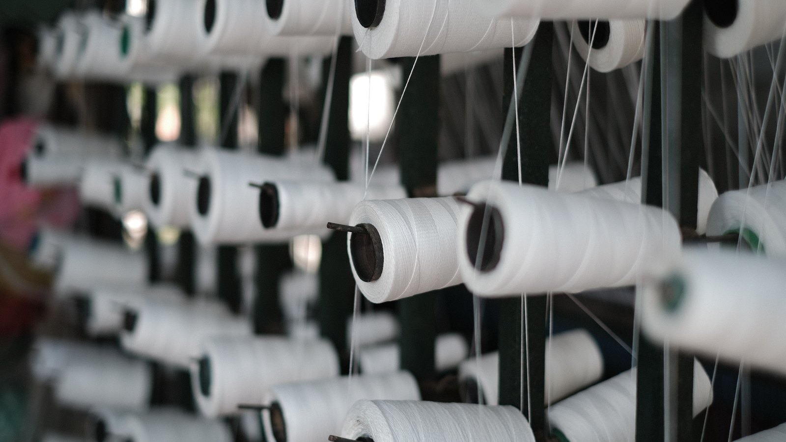 Rows of white yarn bobbins on the steel spindles of a spinning frame, threads running away from each toward the top of the frame.