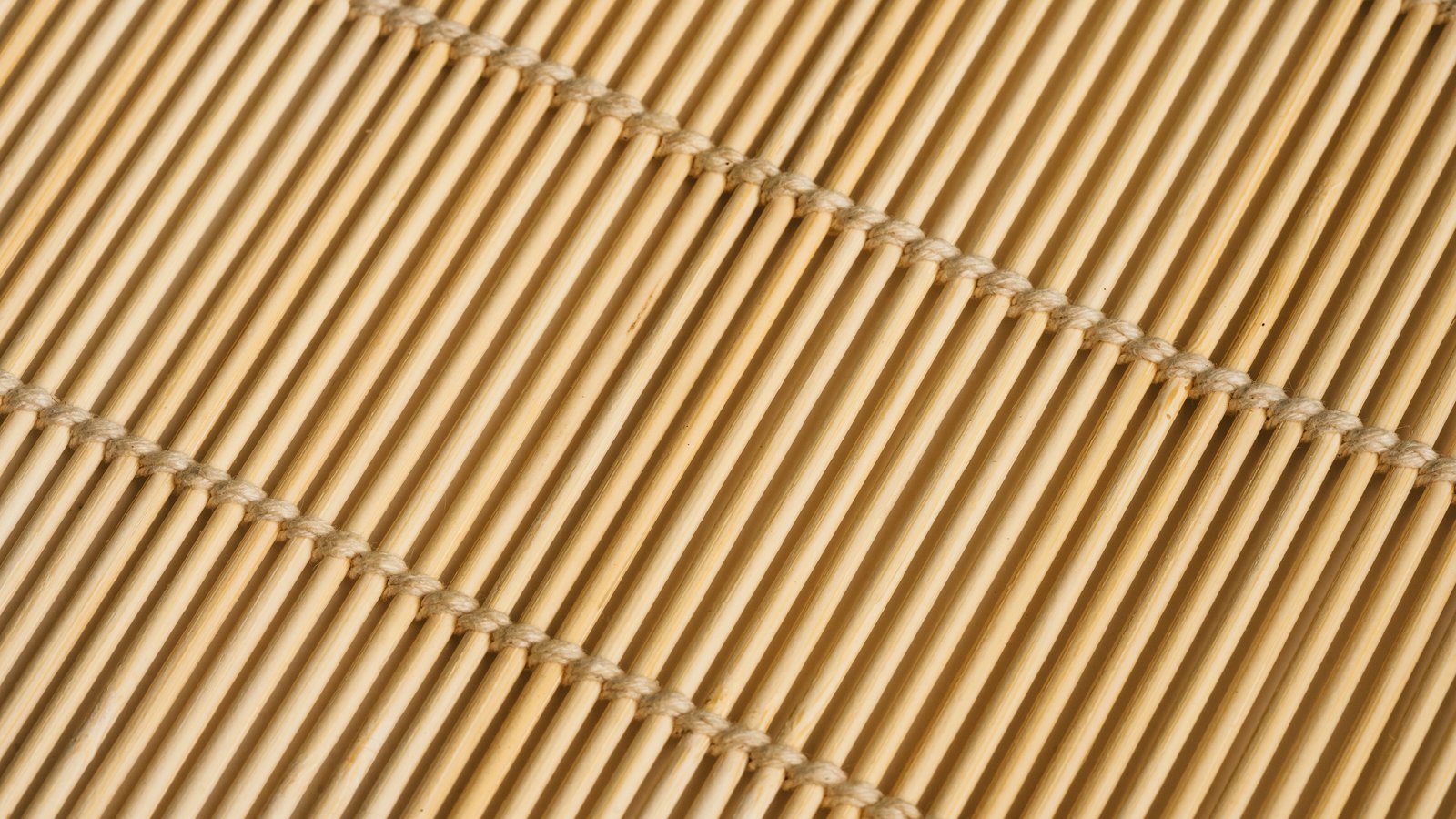 A close diagonal view of a mat of fine pale bamboo slats lying side by side, bound at intervals by twine stitched across them. The parallel slats and the two binding lines fill the whole frame. No text, brand mark, person or place is in view.