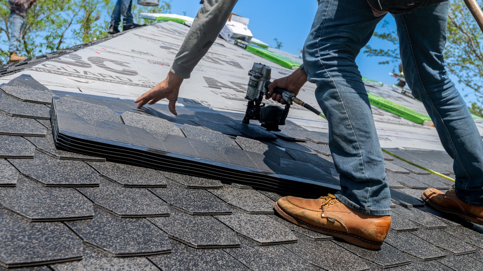 A worker installing shingles on a residential roof with a nail gun under a blue sky.