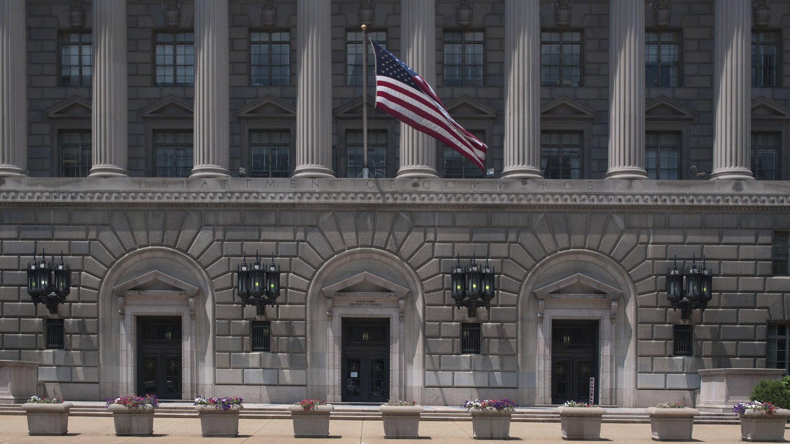 The entrance front of the Herbert C. Hoover Building in Washington, headquarters of the United States Department of Commerce. Three arched stone doorways sit beneath a limestone frieze carved DEPARTMENT OF COMMERCE, flanked by iron lanterns, with a row of fluted columns and an American flag on a staff above, and planters of flowers along the pavement in front.