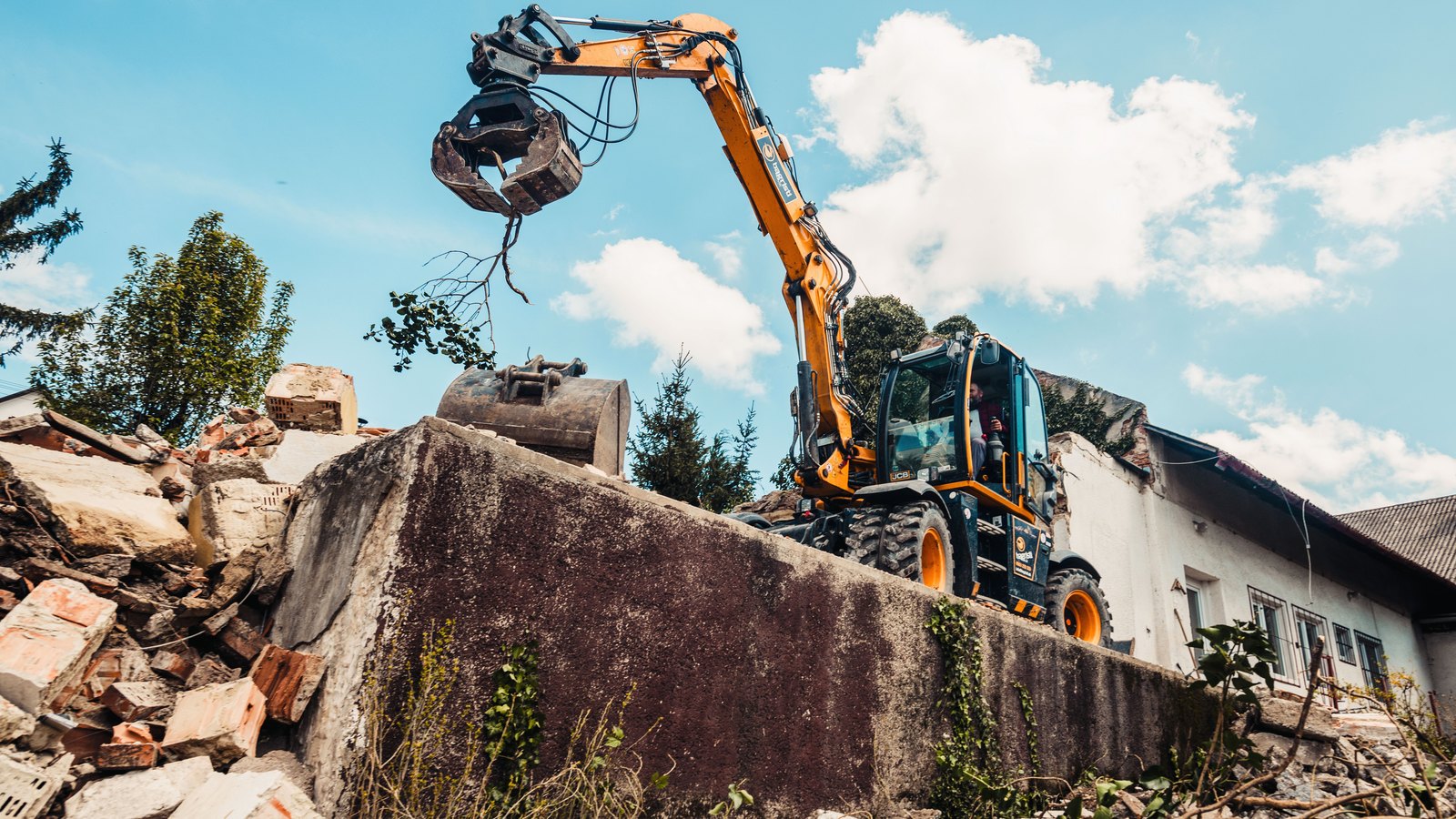 An orange wheeled excavator fitted with a grapple attachment stands on a mound of broken concrete and brick rubble under a bright blue sky with scattered cloud, its arm raised and holding a small clump of leaves, with a plain white single-storey building and evergreen trees behind it. No signage, lettering or place name appears anywhere in the frame. A generic demolition illustration, not a photograph of City of Palms Park or of any property connected to this story.