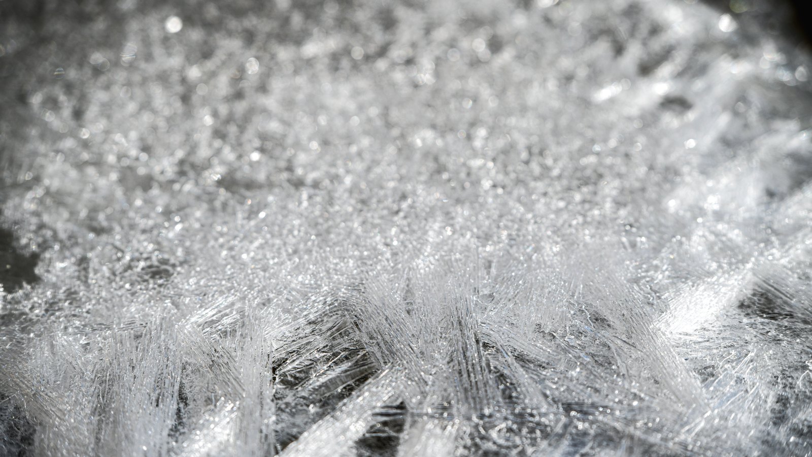 A close photograph of a frozen surface filling the whole frame, thousands of interlocking clear ice crystals catching a hard light so that their sharpest edges flare white against dark shadows underneath. No text, markings, brand marks, people, buildings or objects are in view. A generic material close-up, not a photograph of any exchange, product, filing or party.