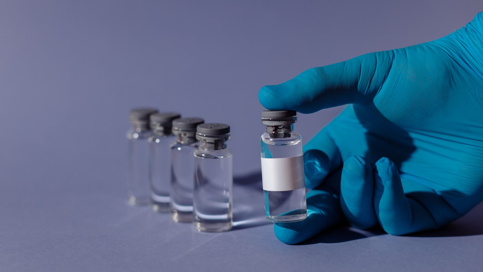 A gloved hand holds an unlabelled medicine vial beside four more vials.