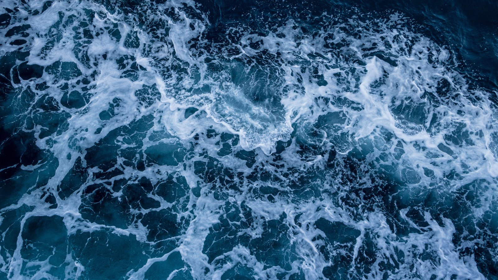 An overhead view of dark blue green seawater churning across the whole frame, threaded with ropes and torn patches of white foam, with no shoreline, horizon, people, boats, buildings, signs or text in view. A generic illustration, not a photograph of a Lee County beach.