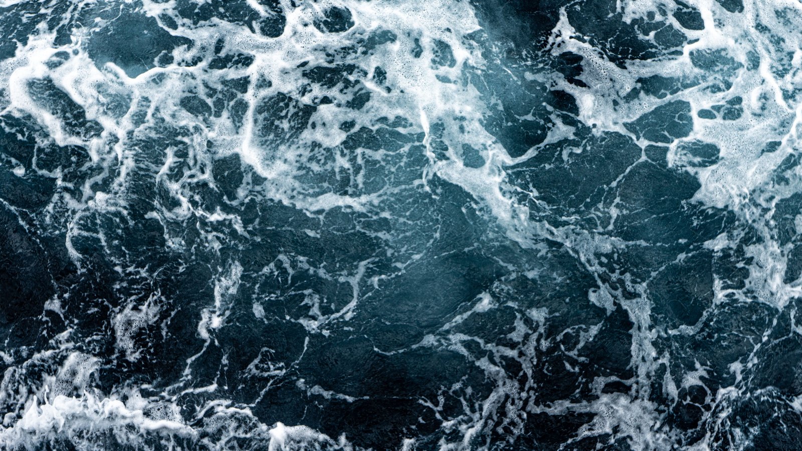A close overhead photograph of churning dark blue green seawater, the surface broken into a web of white foam streaks and bubbles with darker water showing through between them. No shoreline, people, boats, buildings or lettering is in the frame. An illustration of turbulent surf, not a photograph of a Lee County beach.