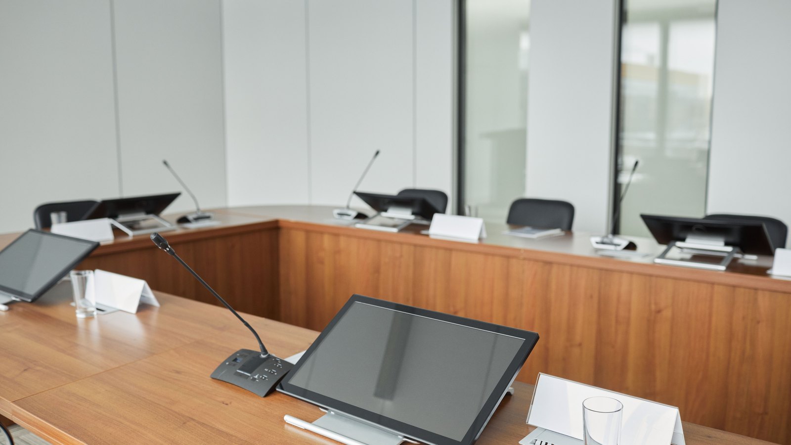 An empty committee room, curved wooden desks facing each other with gooseneck microphones, blank folded name cards and flat monitors at each place.