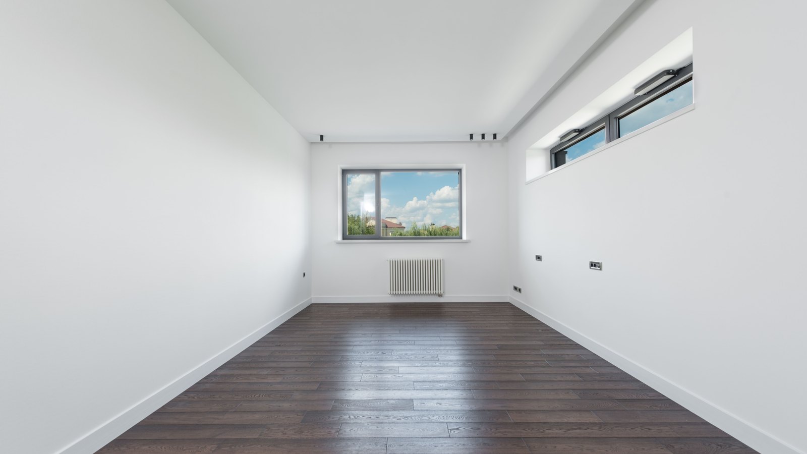 An empty apartment room with bare white walls and a dark wood floor, a single window looking out on trees and rooftops under a bright sky.