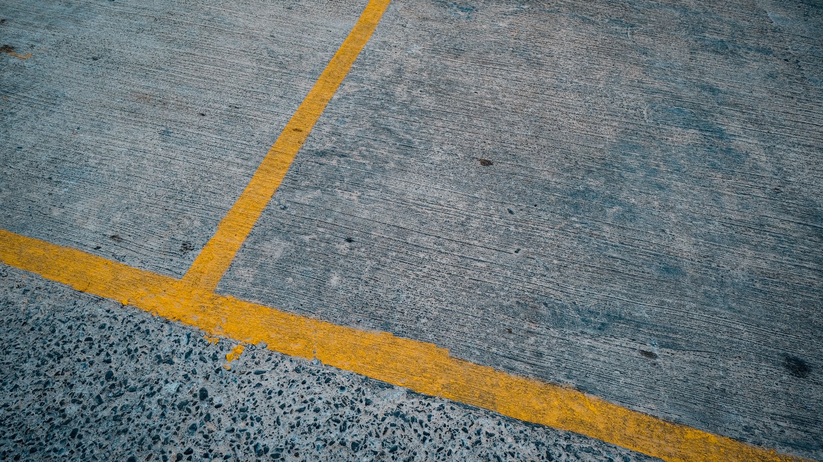 A close overhead crop of worn yellow painted bay markings meeting at a right angle on grey concrete paving, with a band of coarser aggregate along one edge. No vehicle, person, signage or lettering is in view. A generic illustration of a parking bay, not a photograph of the Radio Road transit facility.
