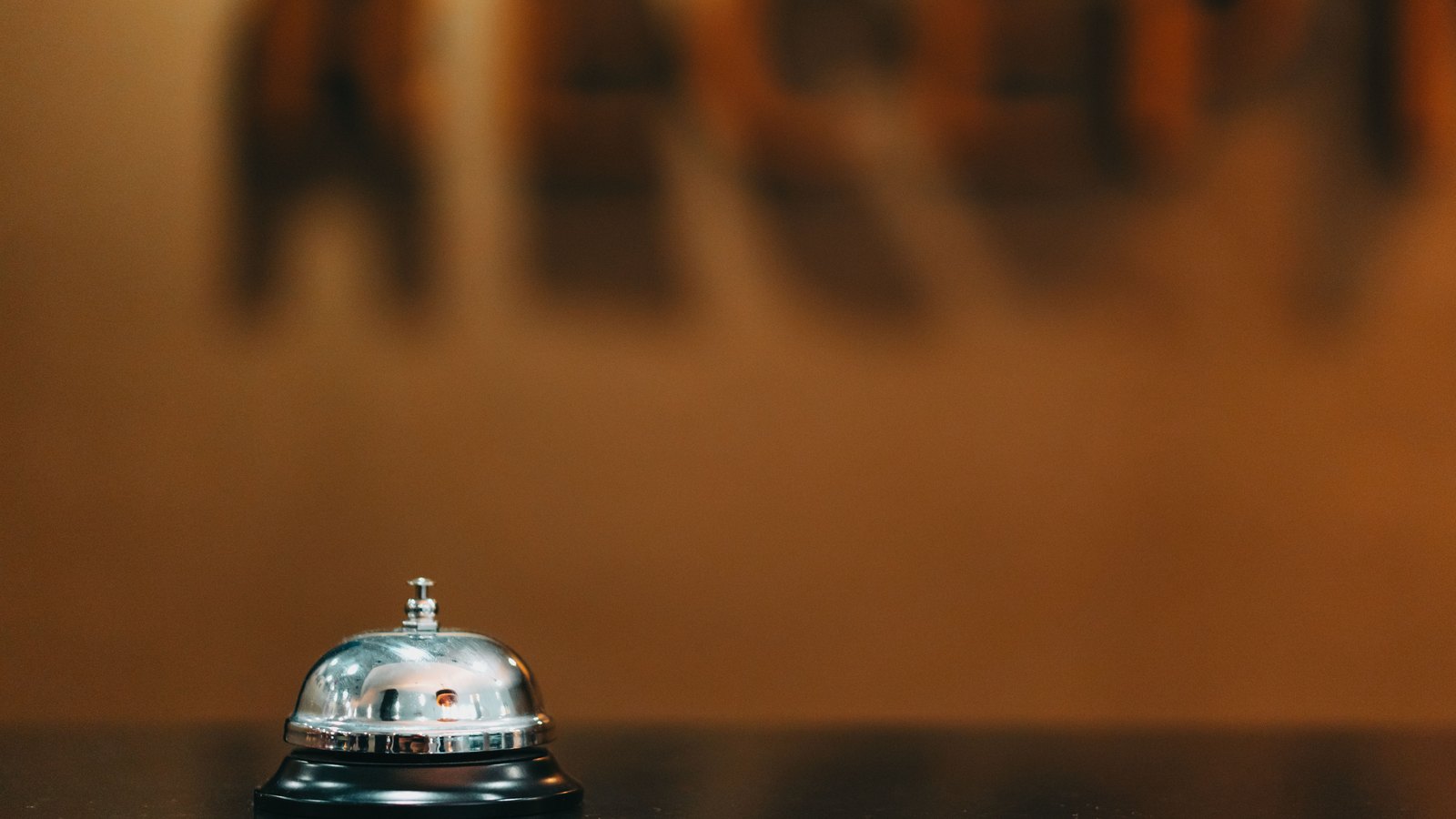 A silver call bell sitting alone at the left of a dark counter, lit warmly against a plain amber wall. Large lettering on the wall behind is blurred past reading and no word can be made out. No person, no sign and no place are identifiable. A generic lodging photograph, not a picture of any Collier County hotel or rental.