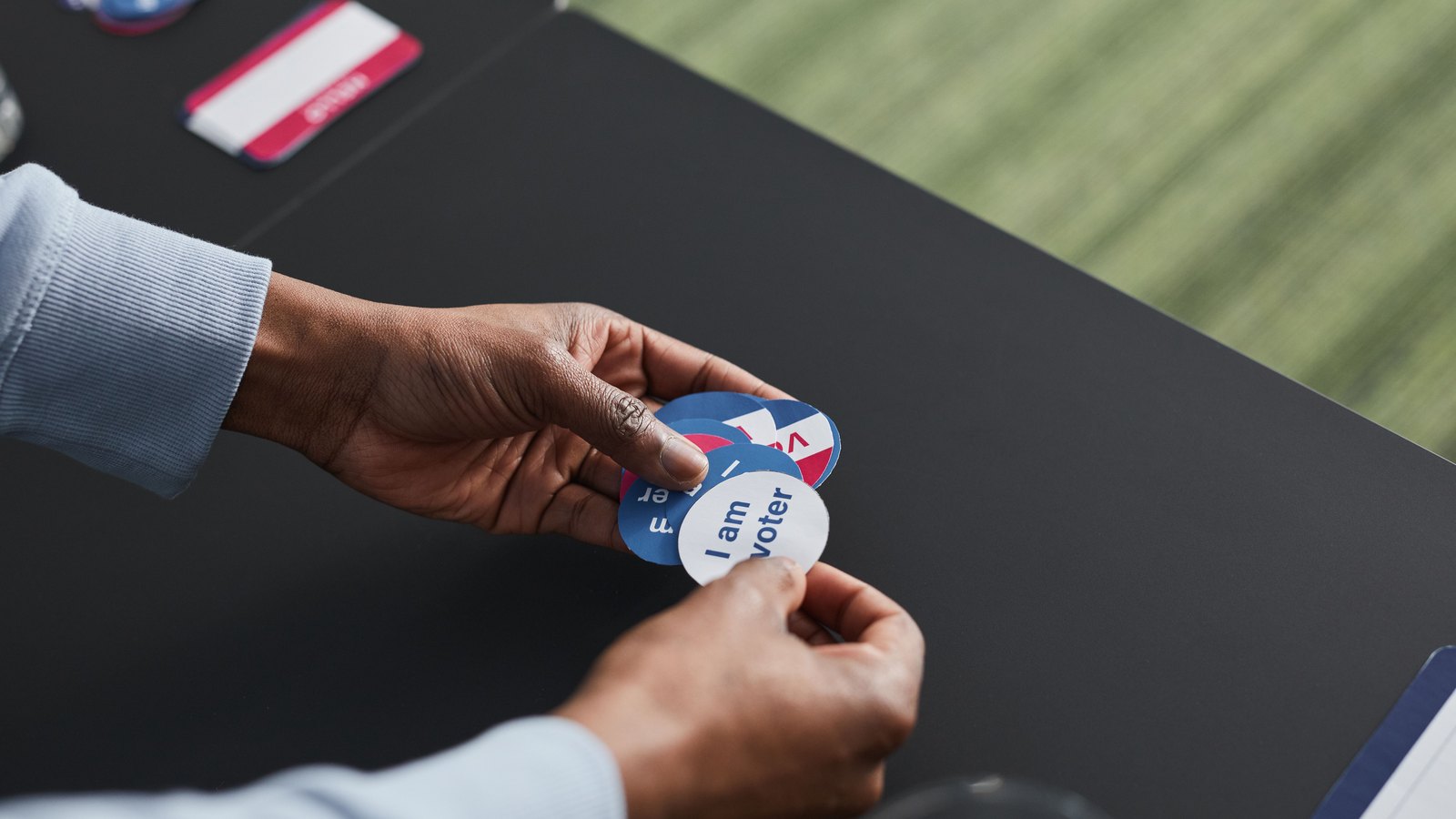 A poll worker at a dark table hands over a small stack of round red, white and blue stickers, the top one reading I am a voter.