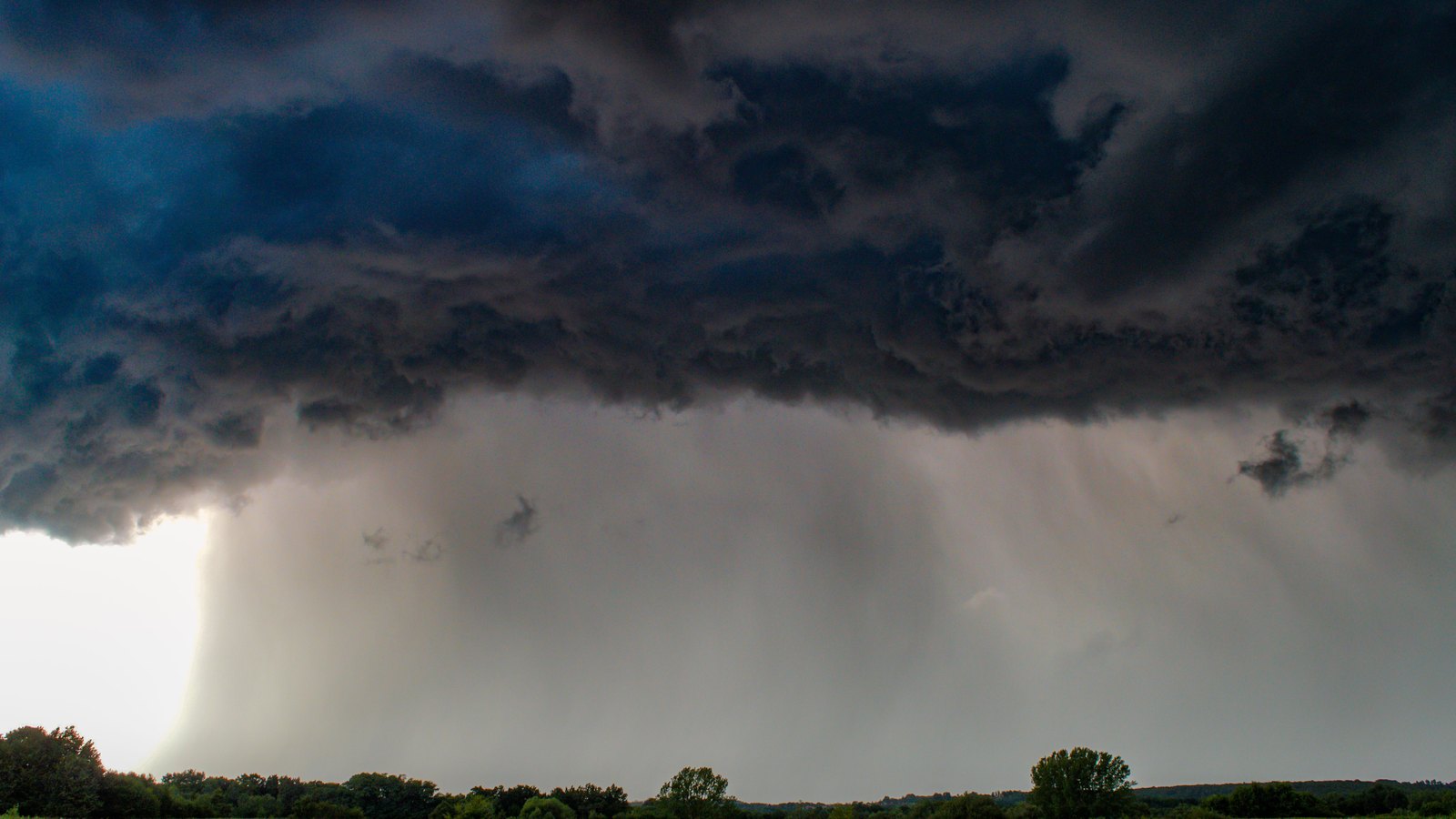 A dark shelf cloud and rain shaft above a flat meadow, not a photograph of this event.