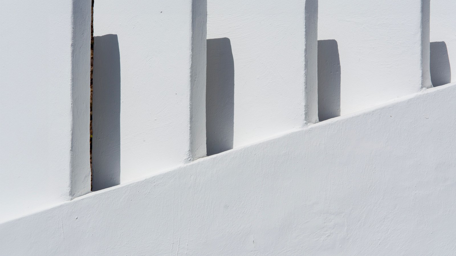 Hard midday sunlight falling across a whitewashed wall, the square posts of a low balustrade throwing short dark shadows onto the render, with no people, signs or text in view.