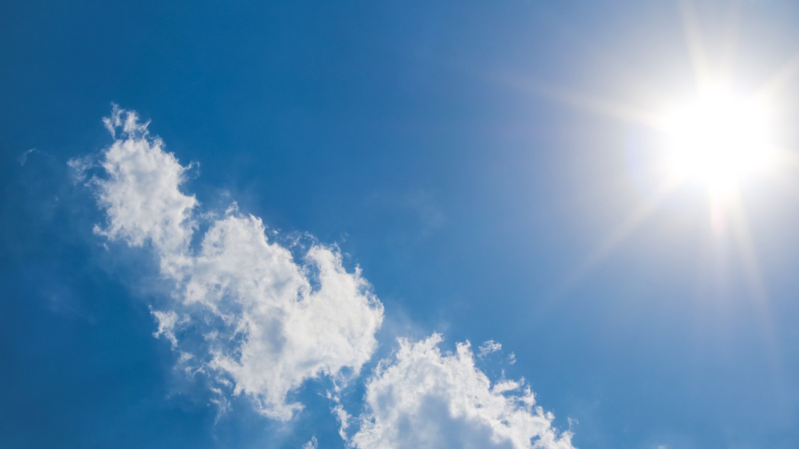 The sun burning white in the upper right of a deep blue sky, throwing a broad flare of glare across the frame, with a diagonal bank of bright white cumulus cloud running across the lower left. No ground, buildings, people, signage or text are in view.