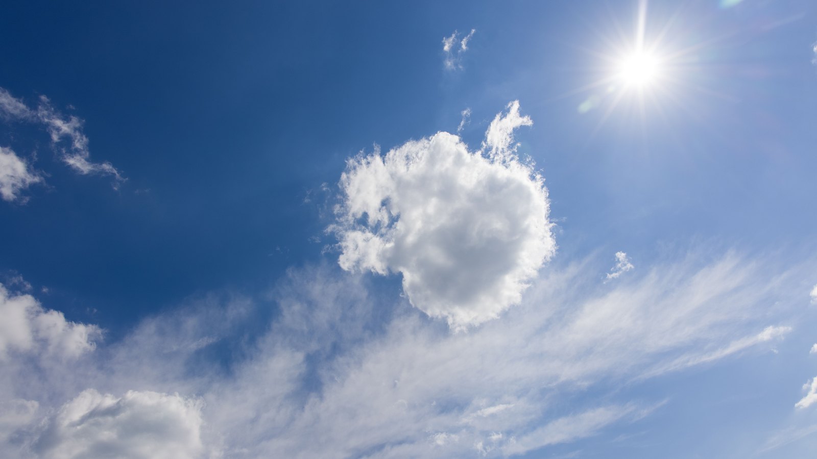 The sun burning high in a deep blue sky, throwing a starburst of glare across thin cirrus and a single bank of white cumulus, with no ground, buildings, people or text in view.