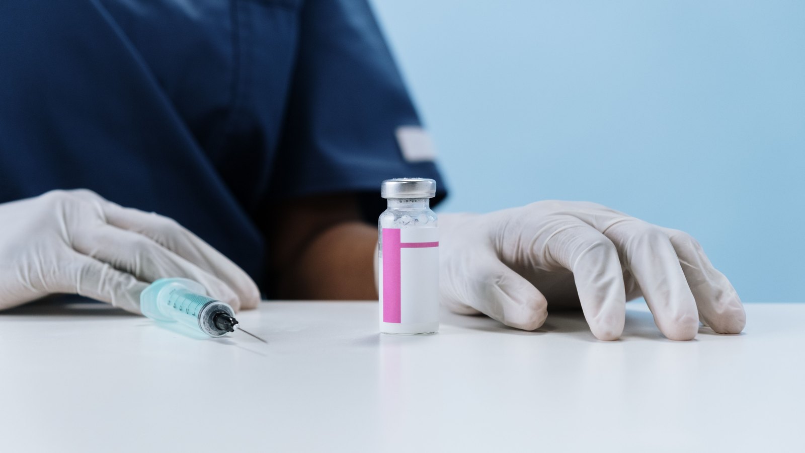 A health worker's gloved hands beside a syringe and an unlabelled vial on a clinic worktop. Stock photo, not a photograph connected to a Collier County case.
