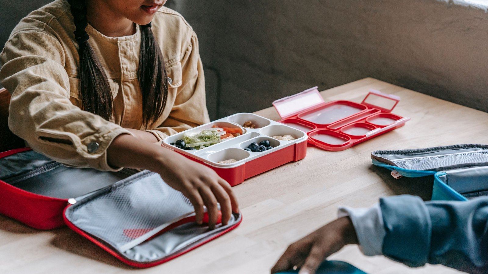 Two schoolchildren at a wooden table with red compartmented lunch trays and insulated lunch bags.