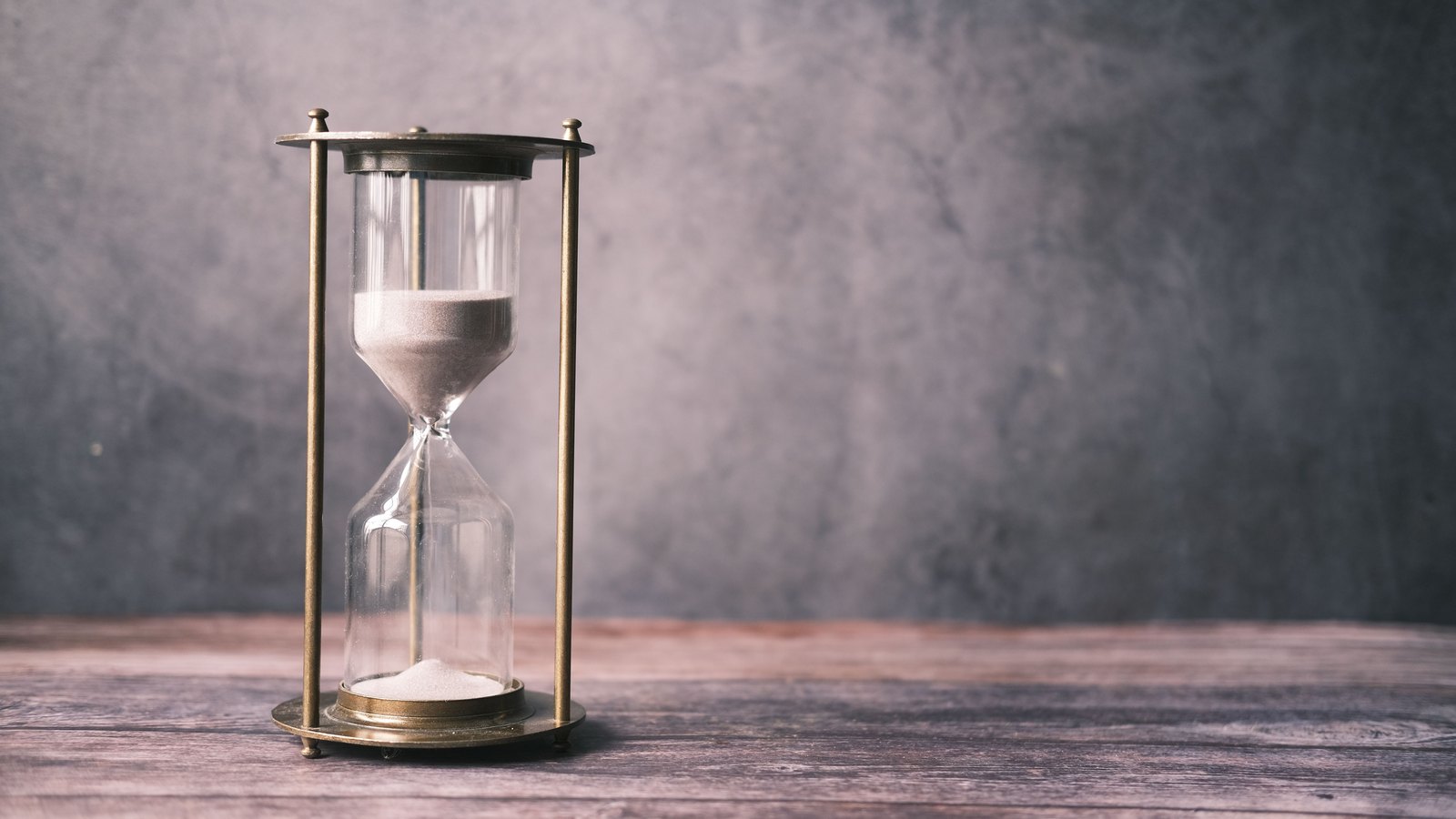 A brass framed hourglass stands on a weathered wooden surface against a plain grey wall, its upper bulb still holding sand while a small heap has gathered in the lower one.