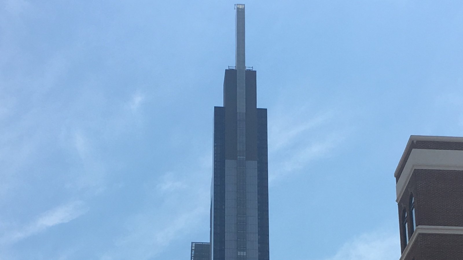The dark glass crown and pale vertical spire of the Comcast Technology Center in Philadelphia against a hazy blue sky, with the corner of a brick building at the right edge.