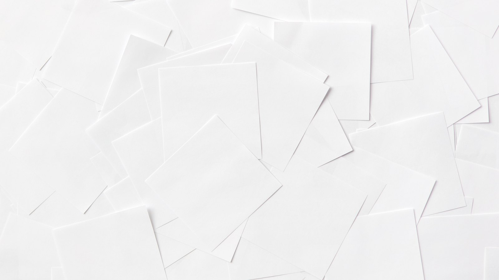 Dozens of blank white sheets of paper lying scattered and overlapping on a white surface, their edges and soft shadows filling the whole frame. Every sheet is empty. No writing, printing, logo, person or place is in view.