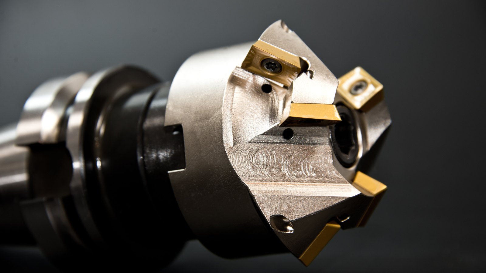 A close view of an indexable milling cutter, its gold-coated carbide inserts bolted around a bright machined steel body against a plain dark background.