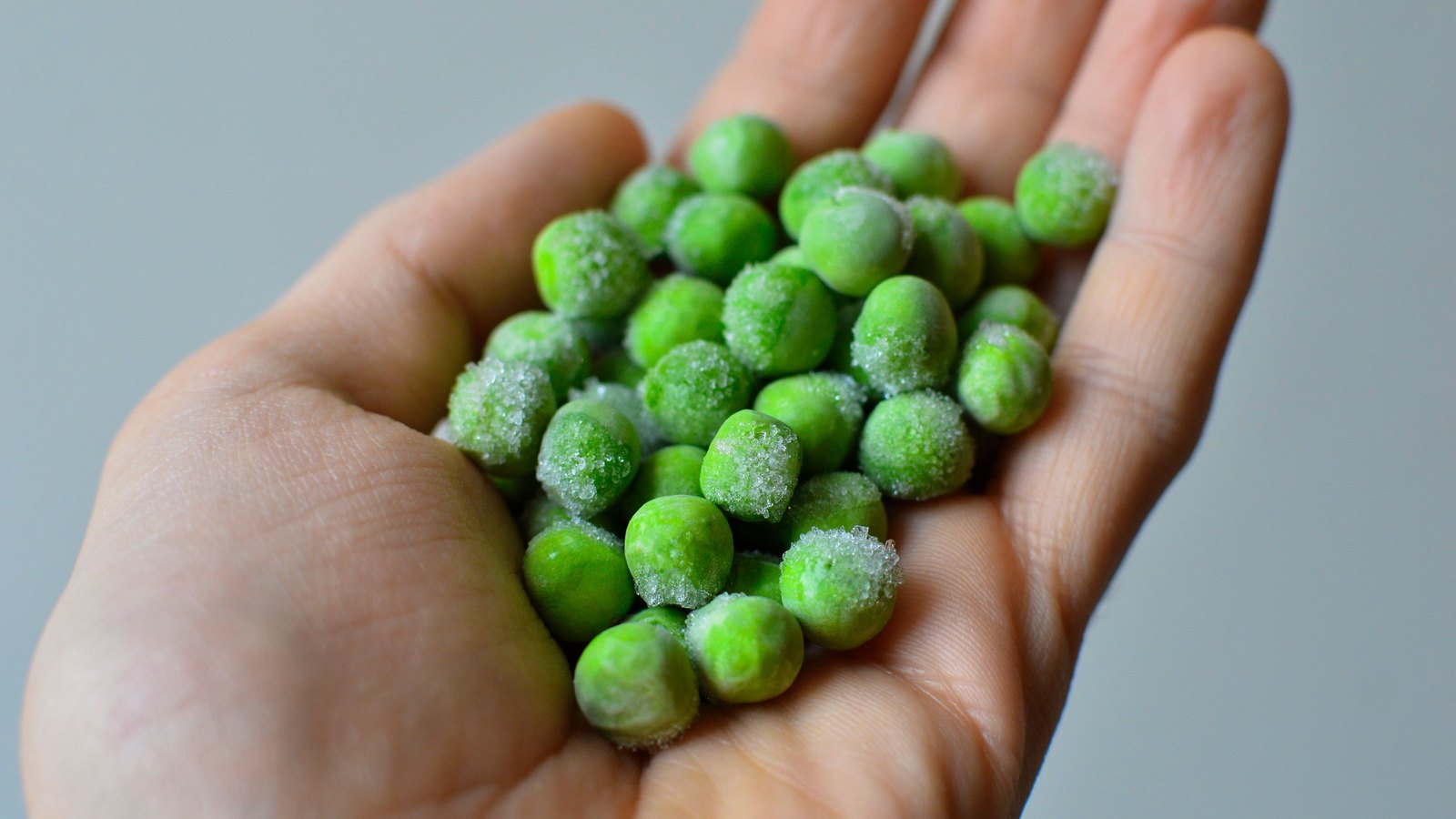An open palm holding a small heap of frozen green peas, ice crystals clinging to them, against a plain grey background. An illustrative photograph, not a product of either company named.