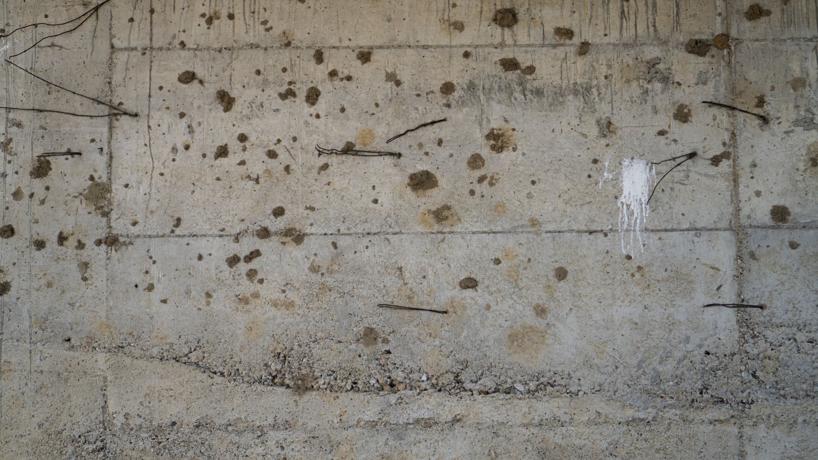 A close photograph of a raw poured concrete wall, filling the frame. Horizontal formwork joints run across it, coarse aggregate is exposed in a band along the bottom, short cut tie wires stand out of the surface at intervals, and there are spatters of dried mud and one run of white paint. No object, person, lettering or brand mark is in the frame.