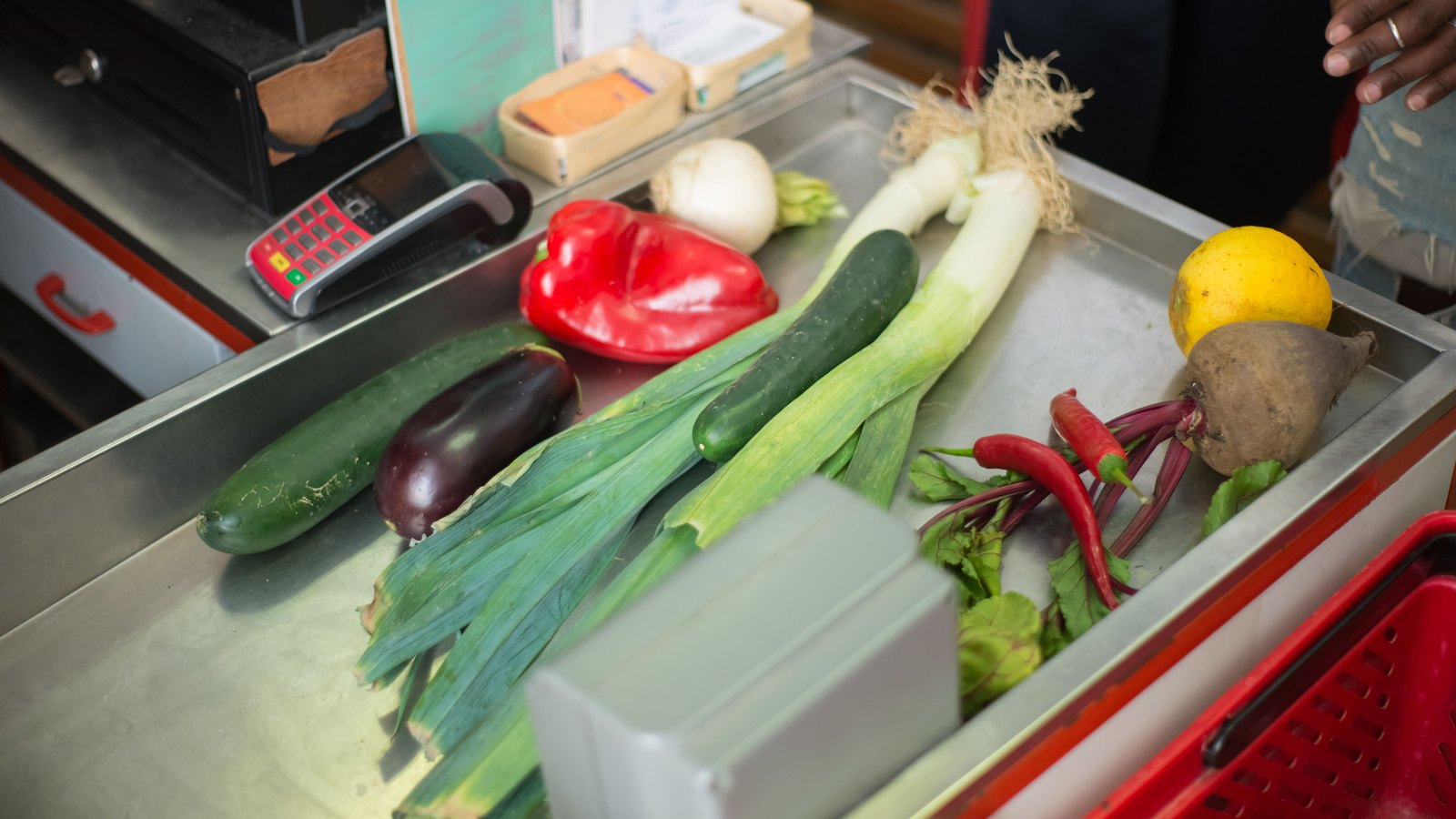 Loose vegetables including a red pepper, leeks, a cucumber, an aubergine, beetroot and chillies laid out along a stainless steel supermarket checkout belt beside a card payment terminal.