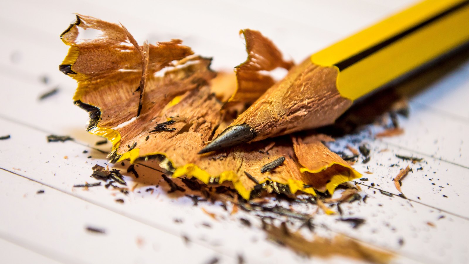 A close view of a freshly sharpened yellow pencil resting on a white surface amid a scatter of curled wood shavings and flecks of graphite.