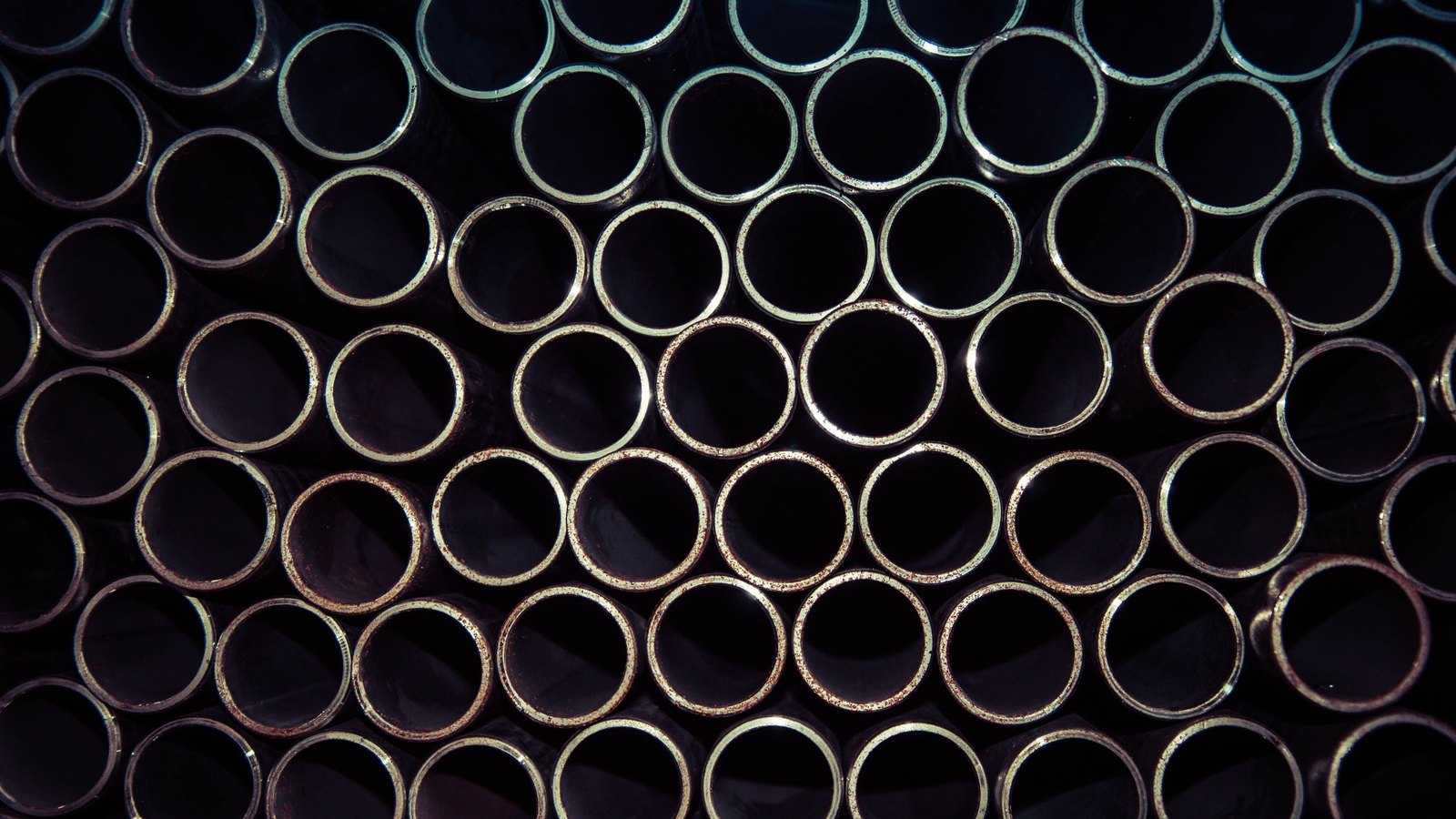 A tightly stacked bank of metal pipes photographed end on, so the frame is filled edge to edge with dark circular openings and bright rims, a few of them freckled with rust. No text, brand mark, person or place is in view.