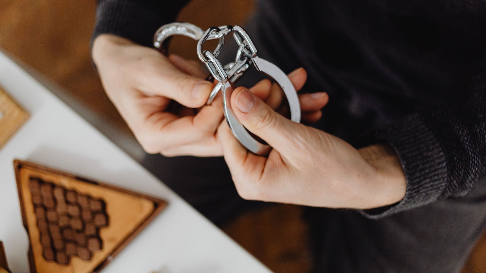 A pair of open steel handcuffs held in two bare hands above a white table, with a wooden puzzle tray and small wooden shapes out of focus below. No face, uniform, badge, agency name, vehicle or location appears in the frame. A stock photograph, not a photograph of this event.