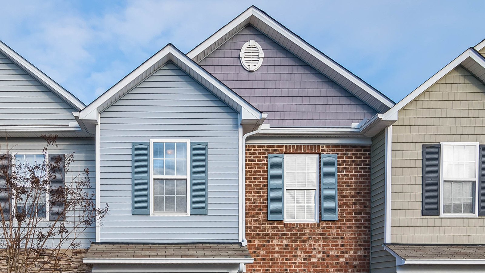 The upper storeys of a row of adjoining American townhouses against a clear blue sky, one clad in pale blue horizontal siding with blue shutters, one faced in red brick, one in beige shingle, each with a white gable and a round louvred vent. No sign, house number, vehicle or person is in the frame.