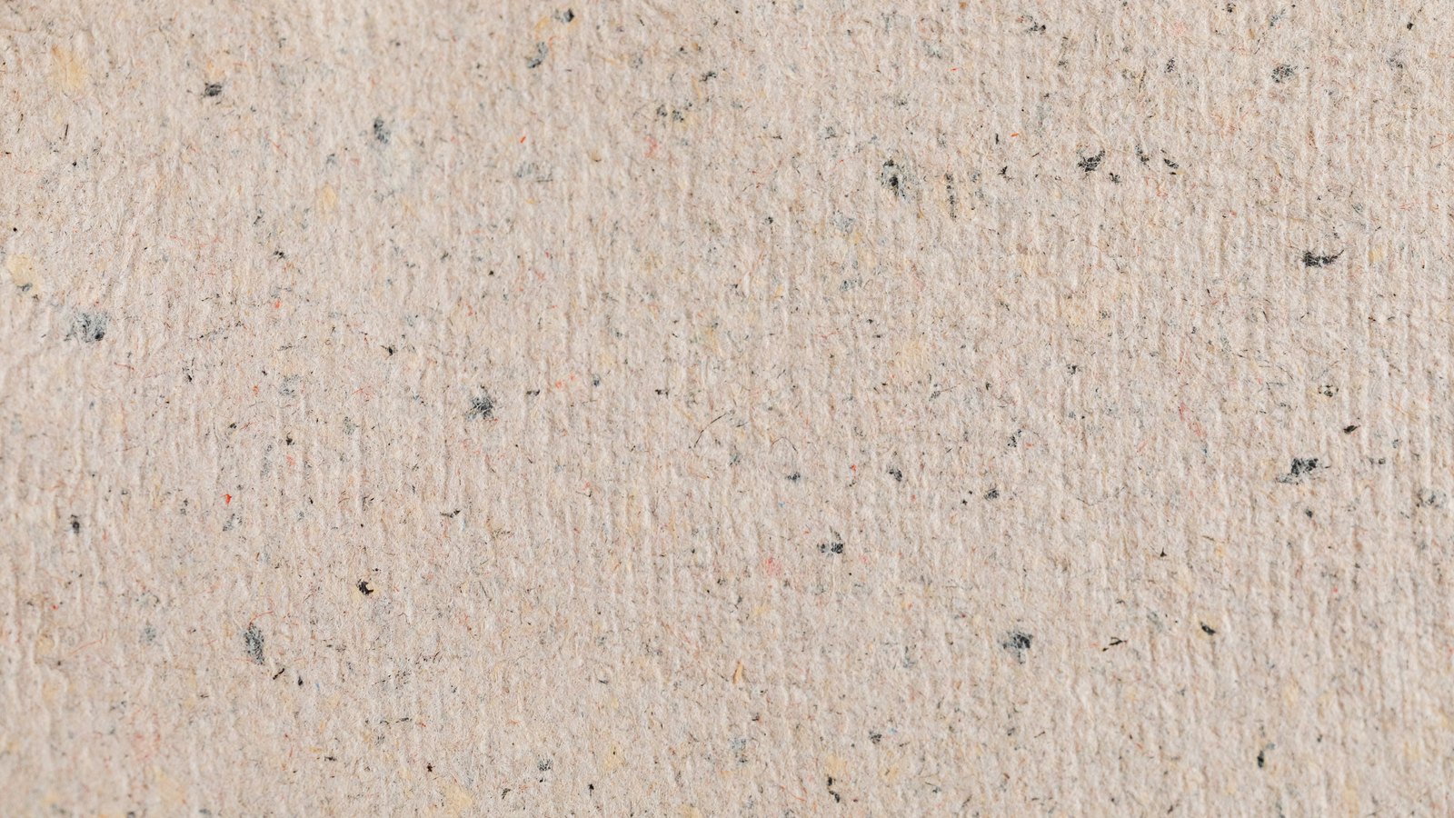 A close photograph of a sheet of pale beige recycled paper, its surface flecked with small dark fibres and occasional specks of red and yellow, the shallow ribbing of the mould running from top to bottom. No object, person, lettering or brand mark is visible.