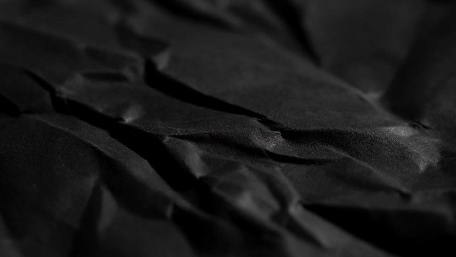 A close macro of heavily crumpled black paper lit from the left, its creases and ridges running diagonally across the frame in low relief.