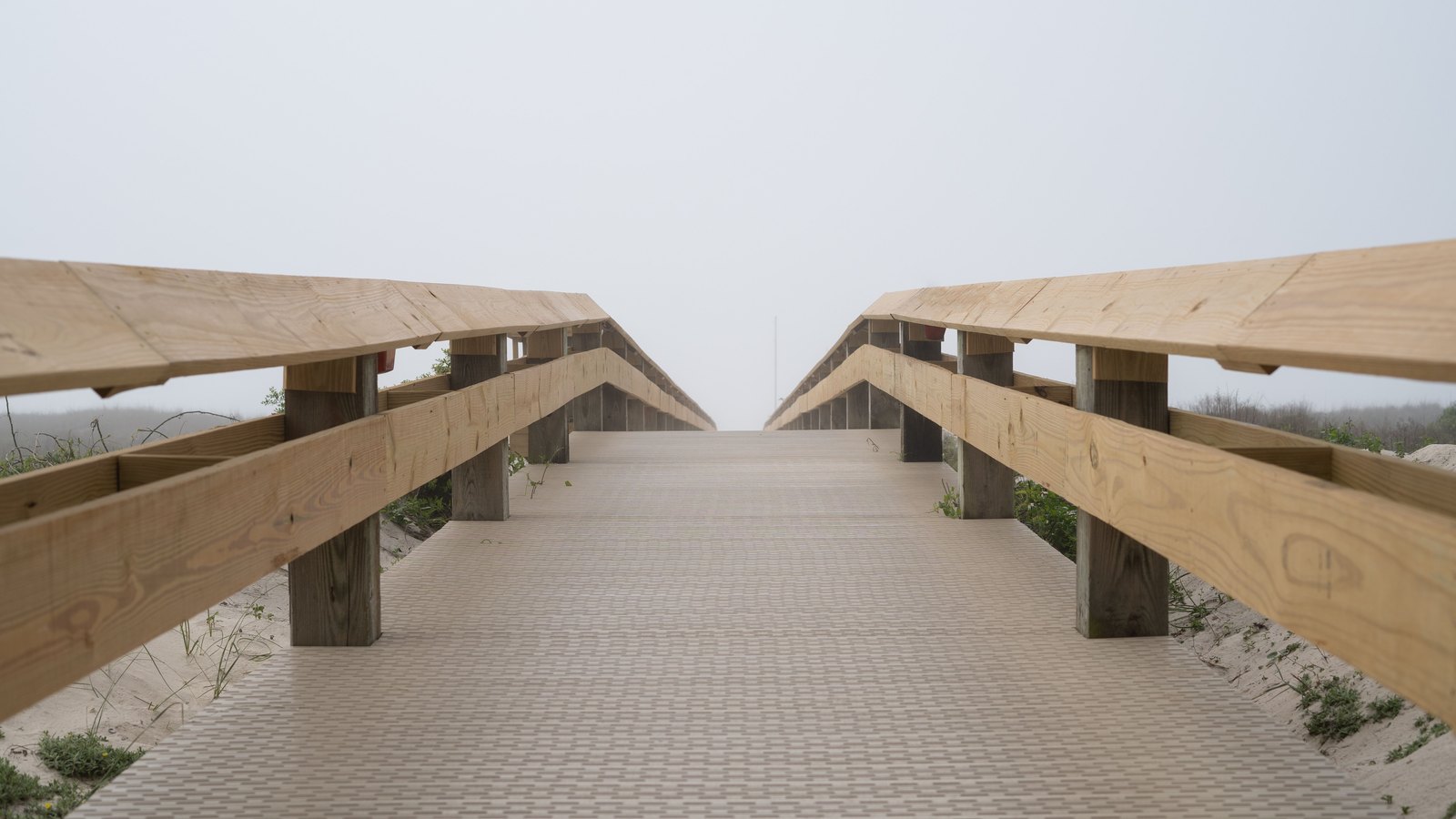 A wooden beach access walkway running away from the camera between pale timber railings, its firm matted surface crossing sand and dune grass toward a flat overcast sky.
