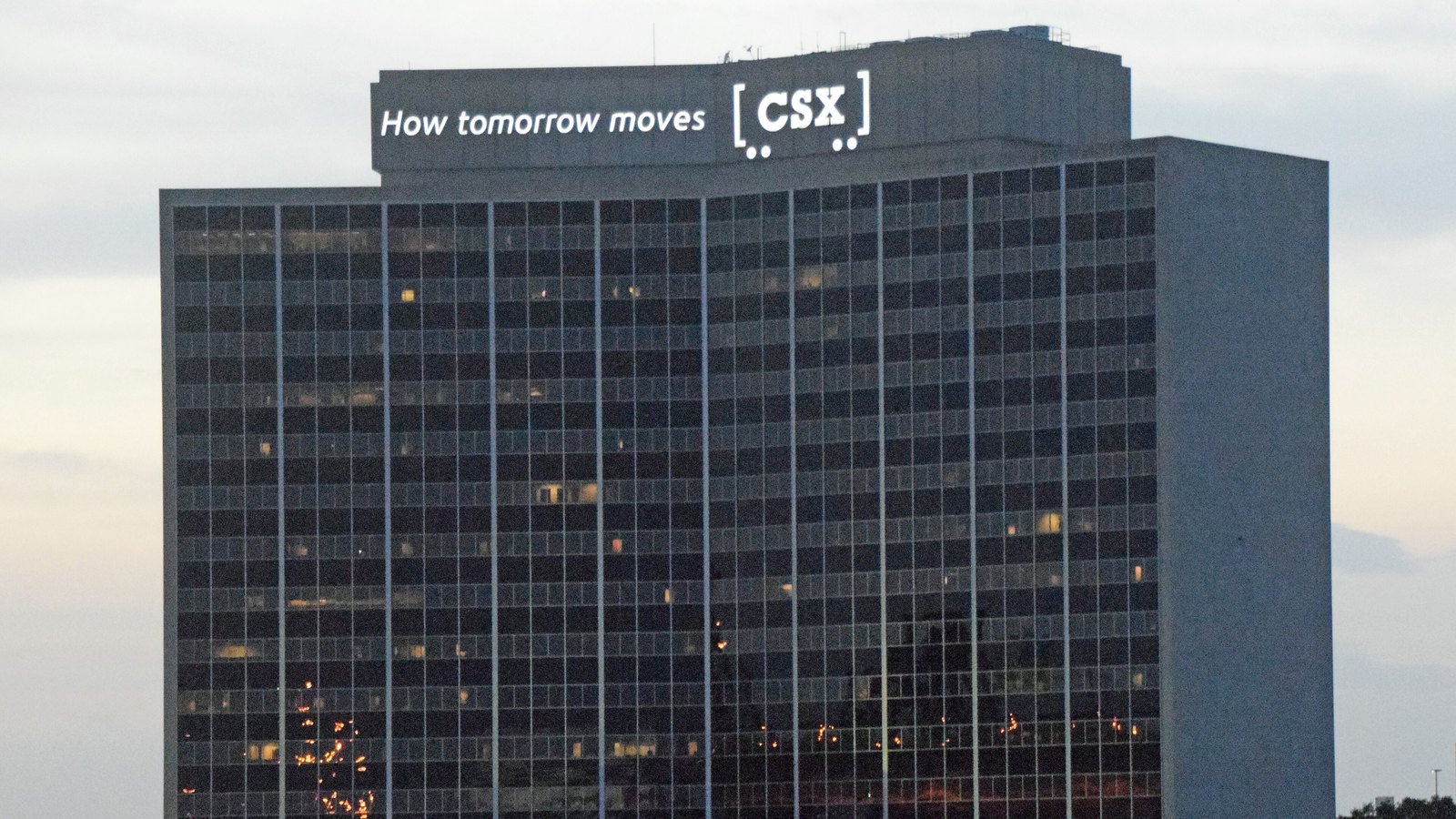 A dark glass office tower at dusk with scattered lit windows, its rooftop crown carrying an illuminated CSX logo and slogan against a pale evening sky.
