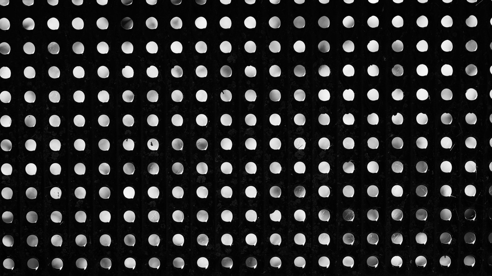 A dark panel drilled with a regular grid of round holes, each one lit from behind so that it reads as a bright disc against black, the rows running edge to edge and filling the whole frame. No text, lettering, brand mark, person or place is in view.