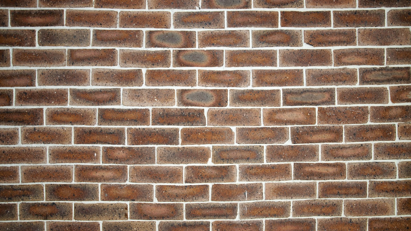 A brick wall photographed square on and filling the frame. The bricks run in regular staggered courses, brown and red with darker scorch marks and pale flecks, separated by narrow white mortar joints. No signage, lettering or fixtures are visible.