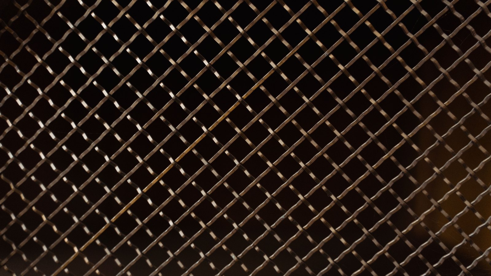 A close photograph of a fine woven metal mesh screen filling the whole frame, the wires crossing on the diagonal in an even grid of small square openings, lit from one side so the bronze coloured strands catch the light against the dark gaps behind them.