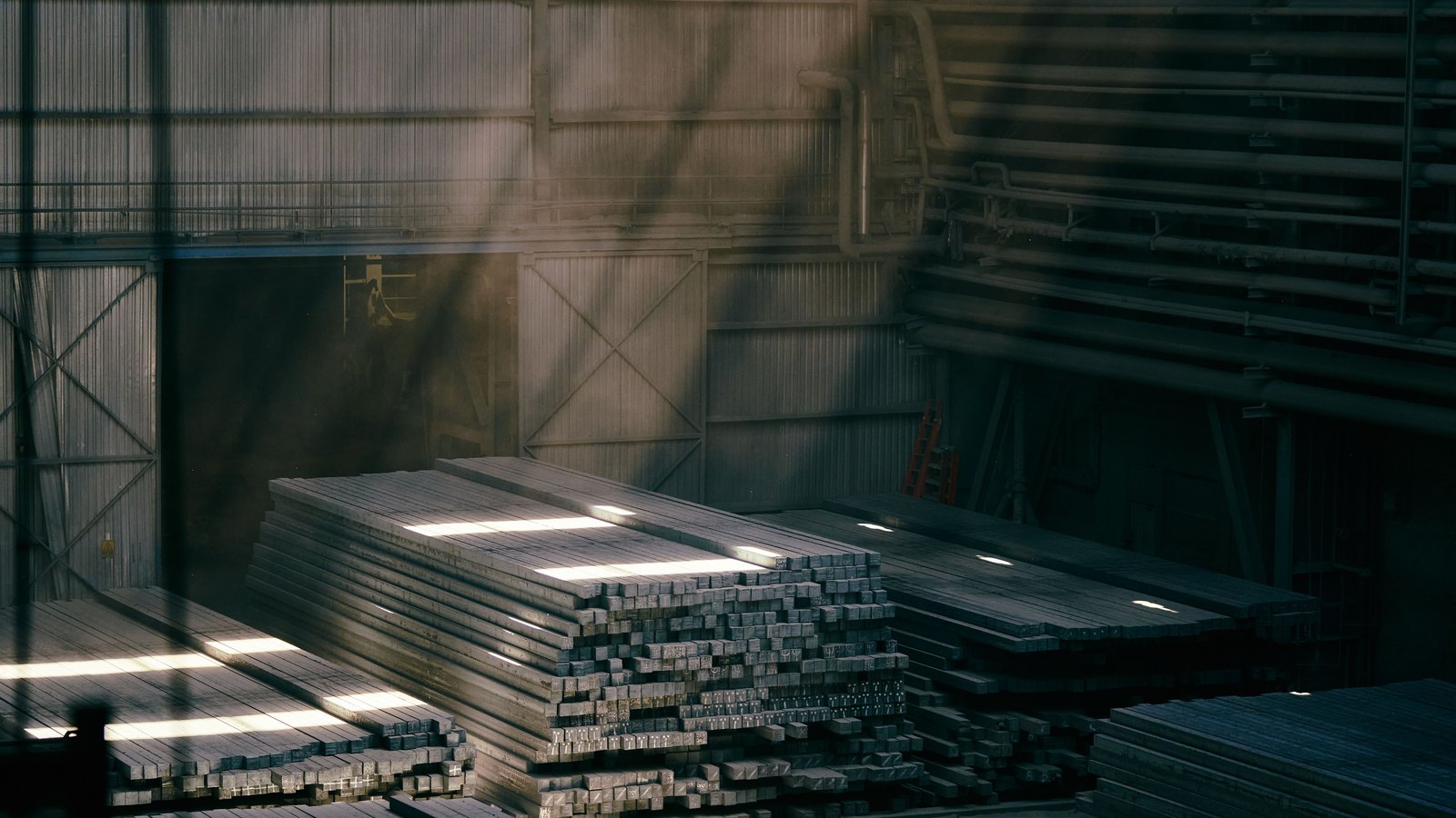 Stacks of square steel bar and flat plate lying on the floor of a mill hall, shafts of daylight crossing them from high windows.