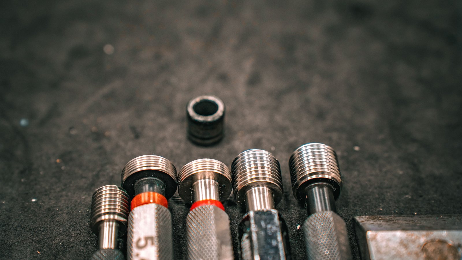 Five knurled steel thread gauges lying in a dark foam tray, their machined threads catching the light against a near black background.