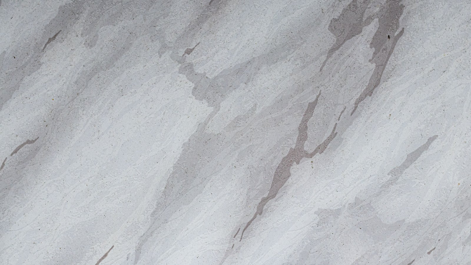 A close photograph of a pale grey surface mottled with softer darker grey streaks running diagonally across it, over a faint woven grain and scattered dark flecks. No people, objects, buildings, signage, text or lettering are visible, and nothing in the frame identifies a place. An abstract surface, not a photograph of any Danaher site or document.