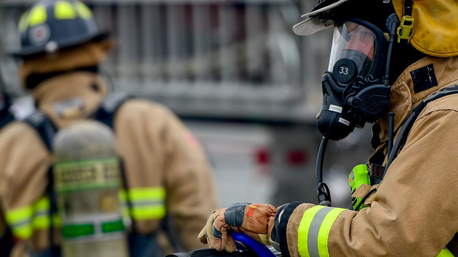A firefighter in breathing gear holds a blue hose line in front of blurred emergency apparatus, used as a general emergency-response illustration and not a photograph of this event.