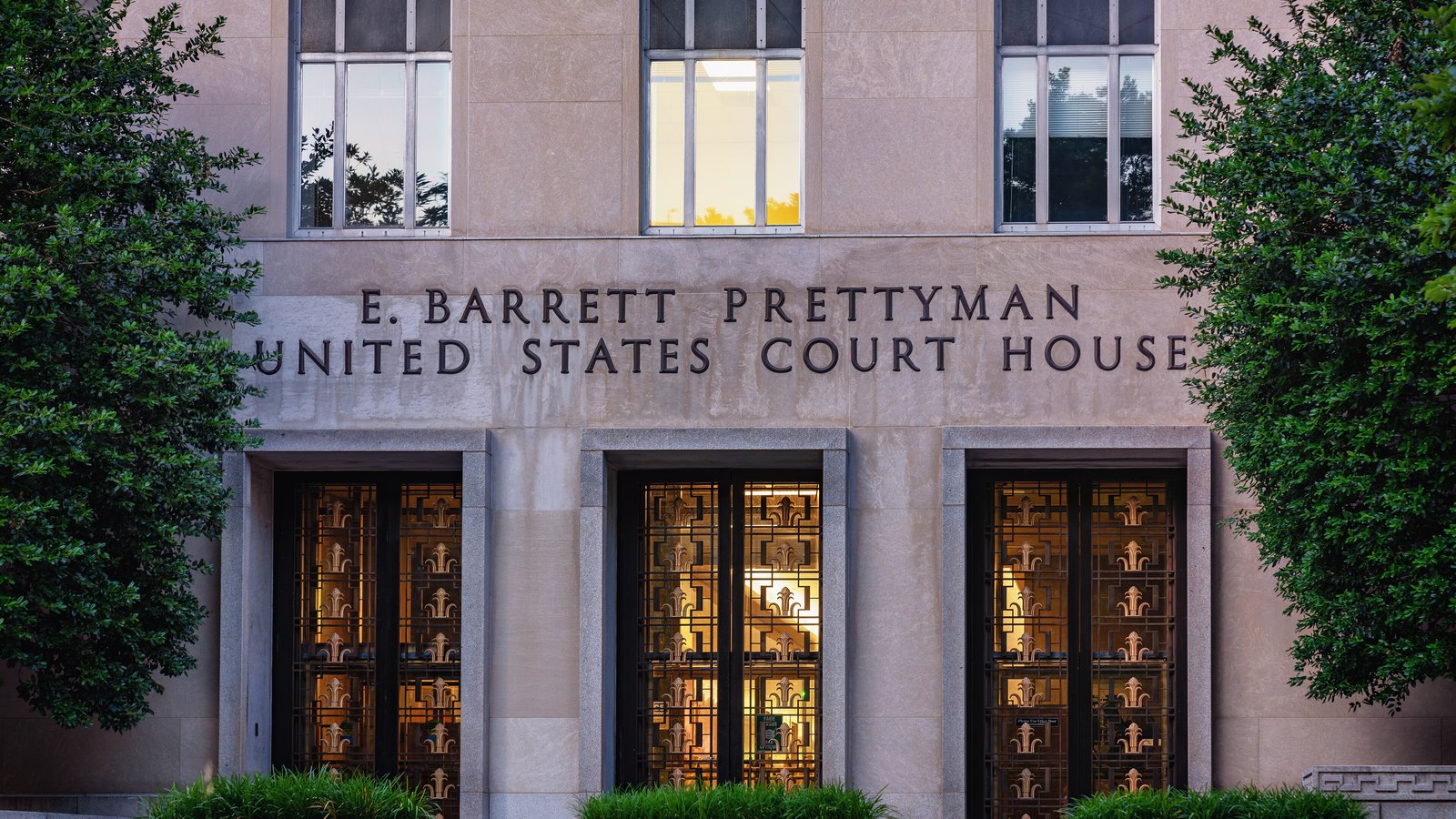 The stone entrance of the E. Barrett Prettyman United States Courthouse in Washington, framed by two trees, with the building name above three bronze doors.