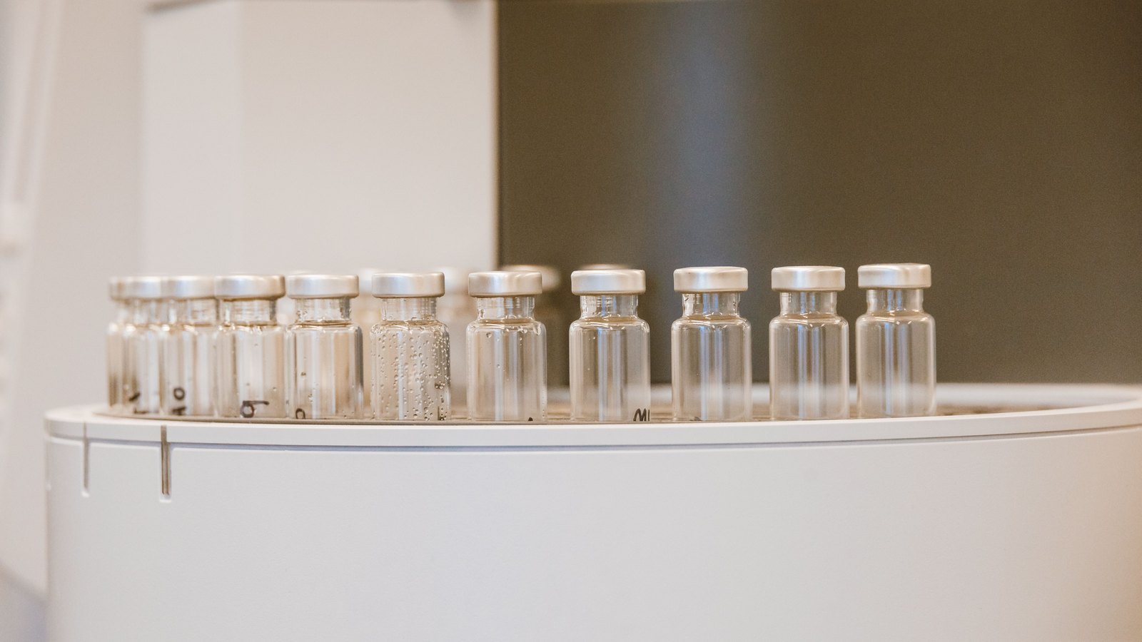 A row of small clear glass vials with silver crimp caps standing upright on a white laboratory carousel, shot at close range with the background thrown out of focus.